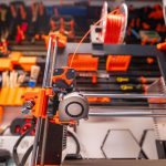 close-up of a 3D printer extruder with orange filament and workshop tools in the background