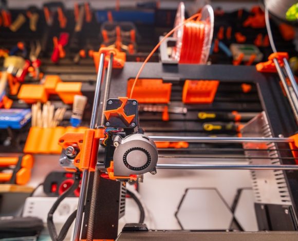 close-up of a 3D printer extruder with orange filament and workshop tools in the background