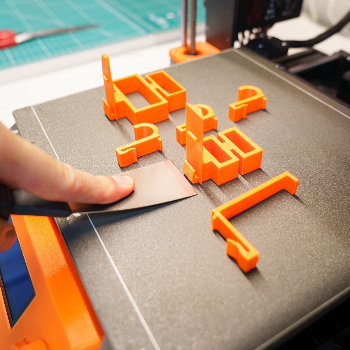 hand using a metal scraper to remove orange 3D printed parts from a print bed product photo