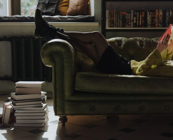 A person relaxing on a green couch reading a physical book with a stack of books nearby, choosing reading over screen time