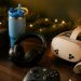 Cozy Christmas gaming desk scene with a Bluetooth speaker, insulated tumbler, wireless gaming headset, controller, and VR headset, framed by string lights and pine branches
