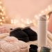 Cozy Christmas scene with black platform slippers, a plush throw blanket, an insulated water bottle, wireless earbuds, and friendship bracelets, framed by a Christmas tree and string lights