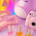 Pastel flat lay of tween girl gifts including a glowing marquee letter A, a Hello Kitty claw clip, a unicorn Squishmallow, a pink Instax camera, and a perfume trio