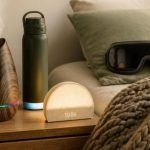 Cozy nightstand wellness scene with an aromatherapy diffuser, smart water bottle, sunrise alarm clock, heated eye massager, and weighted blanket